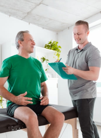 The image is of an older man sitting on a treatment table talking to his physiotherapist